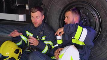 Two tired firefighters are sitting near a fire truck after a fire video