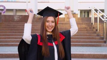 Happy satisfied with her success in study a graduate of the university stands in the mantle of master and expressively swinging a diploma over his head video