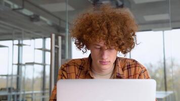 Focused redhead man using laptop sitting in office, serious teenager freelancer distantly working or studying on computer typing online video