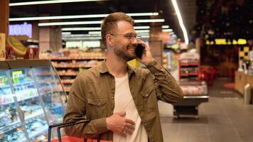 jovem homem com óculos dentro uma supermercado faz compras, escolhe produtos e fala em a telefone, consulta com dele esposa sobre a compras Lista video