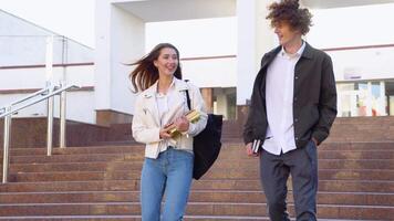 Two stylish students walk near the campus and smile. Positive students friends discuss past lecture near college building video
