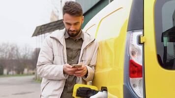 elegant knap mannetje staand in de buurt modern elektrisch auto en sms'en Aan smartphone buitensporig video