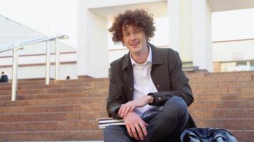 Happy handsome successful student with braces, sits on the steps and smiling, looks into the frame outdoors on the street near the university video