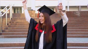 Happy female university graduate stands in a master's mantle and triumphantly waved a diploma over her head video