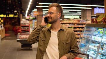 jovem homem dentro uma supermercado fala em a telefone, consulta com dele esposa sobre a compras Lista video