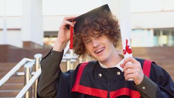 leende röd -hårig lockigt kille med tandställning står i en mästare mantel och innehav på till en hatt och en diplom video