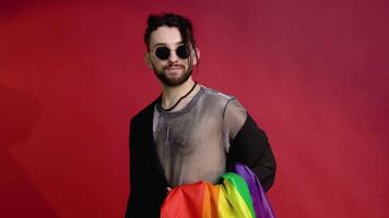 gay uomo vestito nel maglia maglietta e occhiali da sole su rosso sfondo con un' multicolore arcobaleno bandiera. concetto diversità, transessuale, e la libertà video