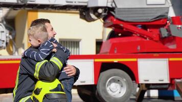 mutig Feuerwehrmann im Uniform Umarmungen ein wenig Junge mit ein Spielzeug gegen ein Feuer Motor video