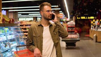 Jeune homme avec des lunettes dans une supermarché fait du achats, choisit des produits et pourparlers sur le téléphone, consulte avec le sien épouse à propos le achats liste video