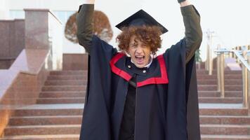 une Jeune frisé marrant diplômé dans maîtrise manteau se réjouit dans compléter le sien études. frisé diplômé avec un appareil dentaire sourit près le Université video