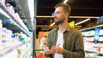 Young man with full shopping basket reading shopping list. A man makes notes in the shopping list video