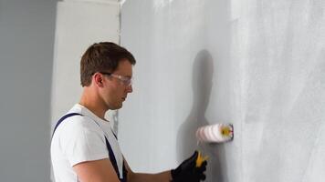Side view of happy young male painter in blue overall and gloves painting wall, with roller in grey color while working on interior design of apartment video