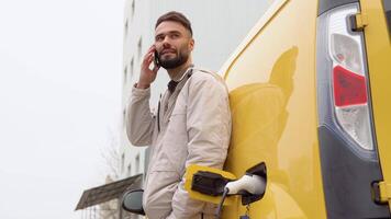 giovane uomo parla su il Telefono mentre il elettrico auto è ricarica all'aperto video