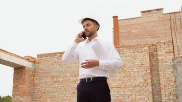 civil ingénieur dans une blanc casque avec les documents dans le sien mains parlant sur le téléphone sur le Contexte de construction. Naturel lumière video