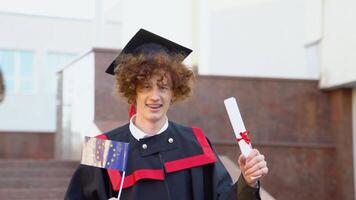 A curly funny graduate in the master's mantle holds a diploma of completion of his studies and the flag of the European Union video