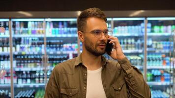 jovem homem anda em dentro uma supermercado faz compras, escolhe produtos e fala em a telefone, consulta com dele esposa sobre a compras Lista video