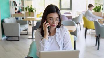 Portrait of happy young office assistant working remotely online sitting with laptop and coffee outside on terrace making notes in agenda in same time speaking with smile on phone with her superior video