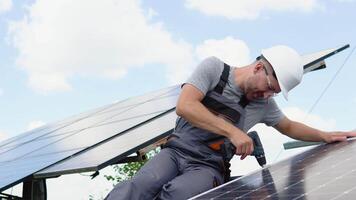 Male engineer in protective helmet installing solar photovoltaic panel system using screwdriver. Electrician mounting blue solar module on roof of modern house video