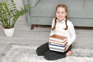 linda pequeño niña sentado en el piso a hogar con un apilar de libros en su manos foto