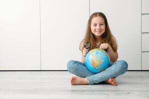 linda pequeño niña sentado en el piso con un globo y un aumentador vaso en su manos alegremente muestra un punto en el globo foto