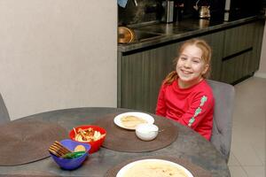 funny kid girl at home at the table in front of a plate of pancakes. photo