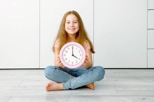 linda pequeño niña sentado en el piso y participación un reloj en su manos ese muestra exactamente 4 4 en punto foto