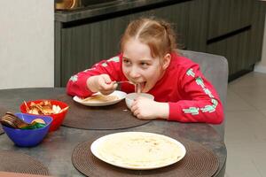 contento pequeño niña a hogar en el cocina a el mesa comiendo condensado Leche con panqueques foto