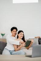 Smiling young couple enjoying a book while relaxing on a sofa in a well-lit room, with a laptop in the foreground. photo