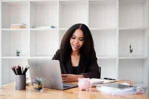 alegre financiero tutor organiza cliente carteras en su computadora portátil, en un moderno oficina con un cerdito banco significando ahorros en el escritorio. foto