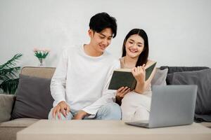 Couple sits comfortably on a couch, sharing a book, with a sense of closeness and a laptop waiting to be used. photo