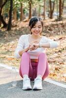 Joyful young woman sitting on a park path, looking at her smartwatch, enjoying a break after a workout session surrounded by nature. photo