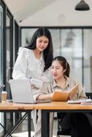 One woman stands guiding another seated at a desk with a laptop, both focused on analyzing a business report in a modern workspace. photo