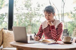 mujer en rojo y blanco a rayas suéter escribe notas junto a su ordenador portátil en un brillante hogar ambiente. foto