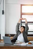 Professional woman stretches with eyes closed, taking a relaxing break at her workspace with a laptop and books. photo