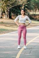 Woman in a white crop top and pink leggings stands with hands on hips on a yellow-lined path, trees in background, wearing a smartwatch. photo