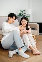 Young man in a white long-sleeve shirt and light blue jeans sits on the floor with a woman in a sleeveless dress, both enjoying content on a smartphone together. photo