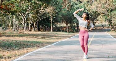 Woman in a white cropped top and pink leggings shielding her eyes from the sun, standing on a sunlit park pathway with trees. photo