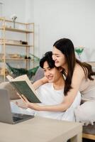 young man in a white t-shirt and a woman in a beige sleeveless dress are engaged in reading a hardcover book together on a sofa. A laptop is open beside them. photo