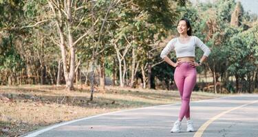 Smiling woman in a white cropped top and pink leggings stands with hands on hips on a paved park pathway, trees in the background. photo