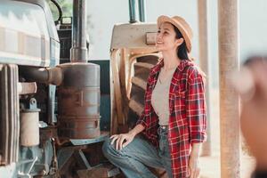 Joyful farmer in a straw hat and plaid shirt leans on a blue tractor, posing with hands on her hips. photo