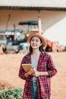 Smiling young farmer holding a notebook stands proudly on her farm with agricultural machinery in the background. photo