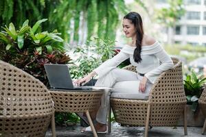 Engaged freelancer leans forward to work on her laptop, seated in an inviting garden setting with lush plants around. photo