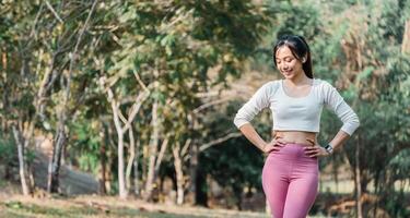 Smiling woman in a white cropped top and pink leggings stands with hands on hips on a paved park pathway, trees in the background. photo