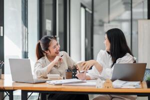 Two Asian businesswomen share a laugh during a coffee break in a modern office setting, with laptops and work materials on their desk. photo