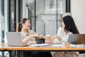 Two joyful businesswomen laughing and chatting over a cup of coffee at a wooden office desk with laptops. photo