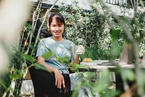 sonriente mujer en un azul camisa participación un vaso, sentado a un mimbre silla en un lozano jardín cafetería. foto