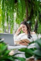 Woman experiences a moment of stress while working on her laptop in a tranquil garden, surrounded by hanging ferns and foliage. photo