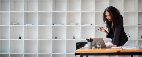 Pensive businesswoman in a dark blazer holds a pen and studies her laptop screen at an office desk. photo