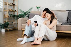 Relaxed and smiling, an Asian couple shares a close moment while looking at a smartphone, sitting on the living room floor. photo