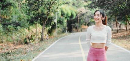 Young woman in a white crop top and pink leggings stands smiling on a sunlit park pathway with greenery in the background. photo